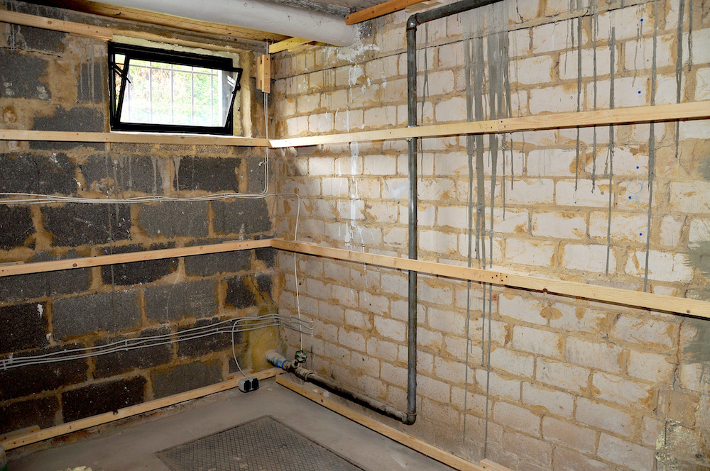 Kellerraum mit freiliegenden Ziegelwänden, Wasserflecken, Holzleisten, kleinem vergittertem Fenster und Rohren an der Wand.